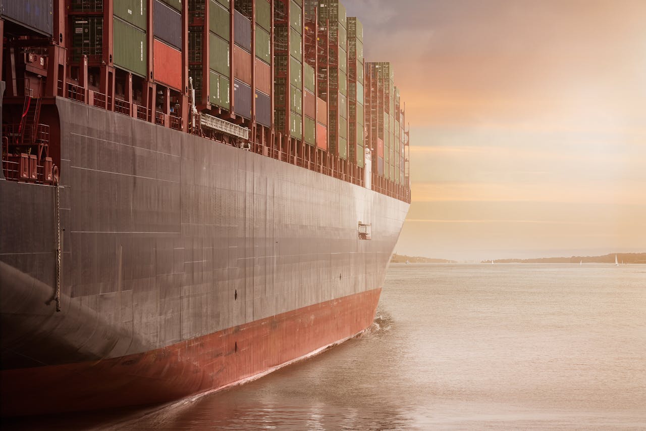 A colossal cargo ship loaded with containers navigates through calm waters against a vibrant sunset sky.